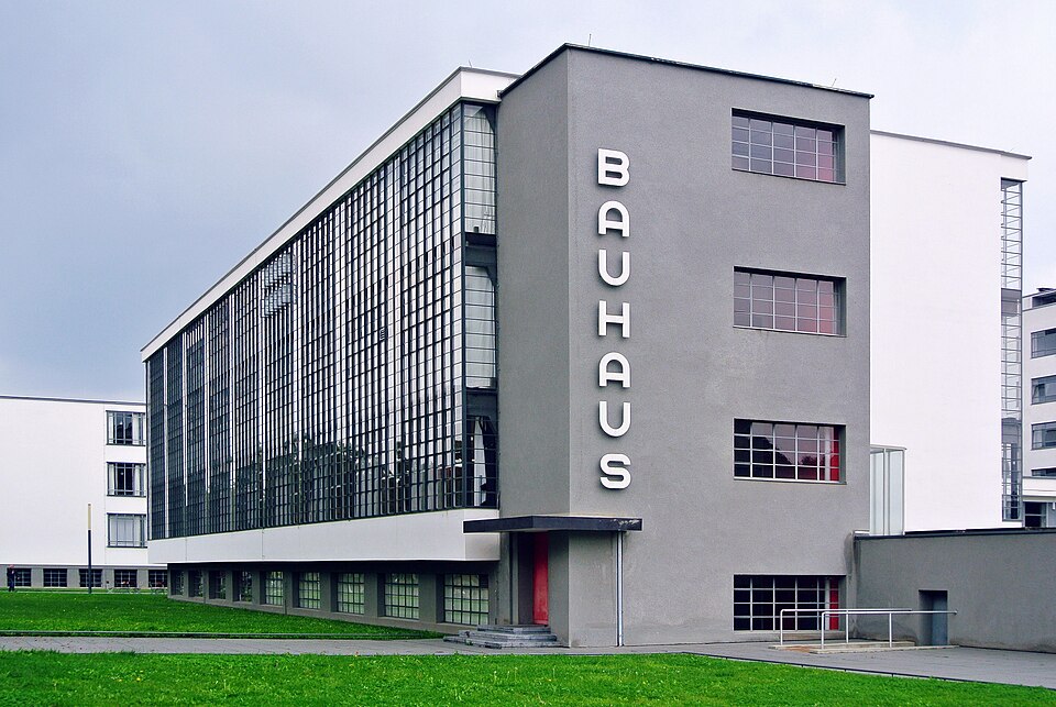 A modernist building with white walls and a large grid of glass windows on multiple stories, set against a clear sky