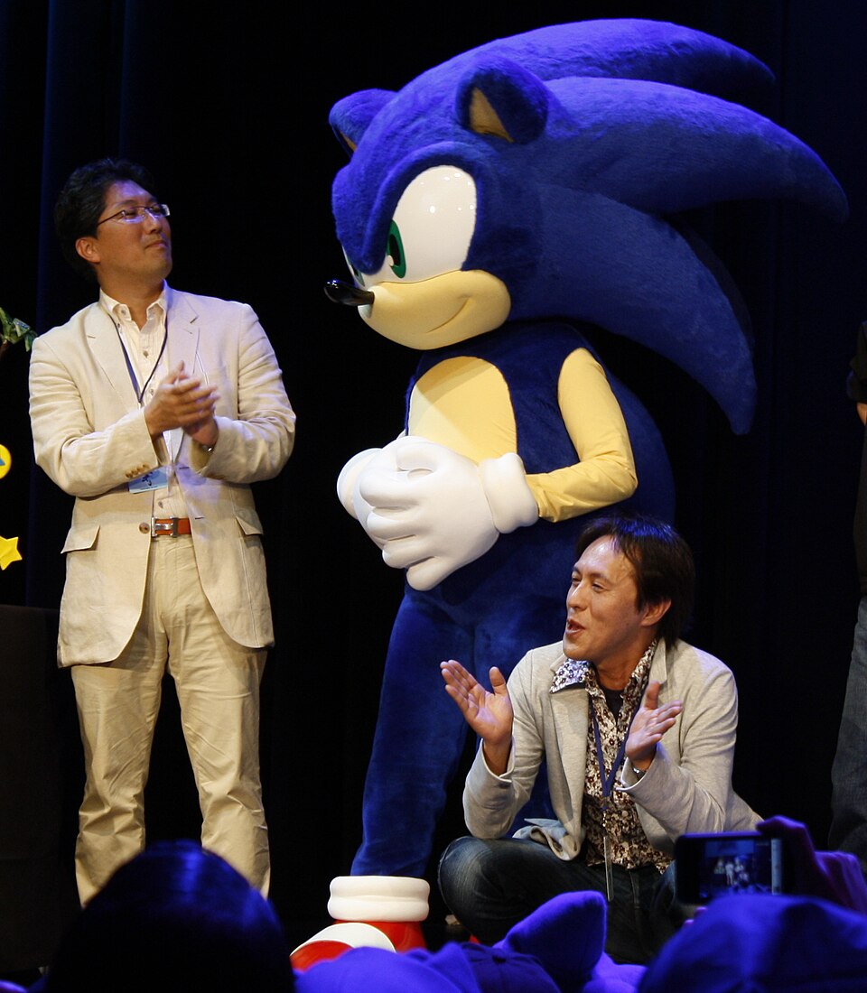Three men flanking a costumed Sonic figure, singing in a crowd